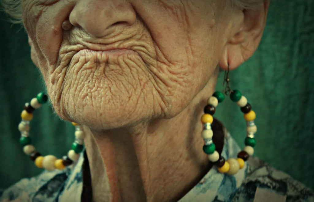 Close-up of an elderly woman's face with detailed wrinkles, wearing large beaded earrings against a green background.