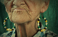 Close-up of an elderly woman's face with detailed wrinkles, wearing large beaded earrings against a green background.